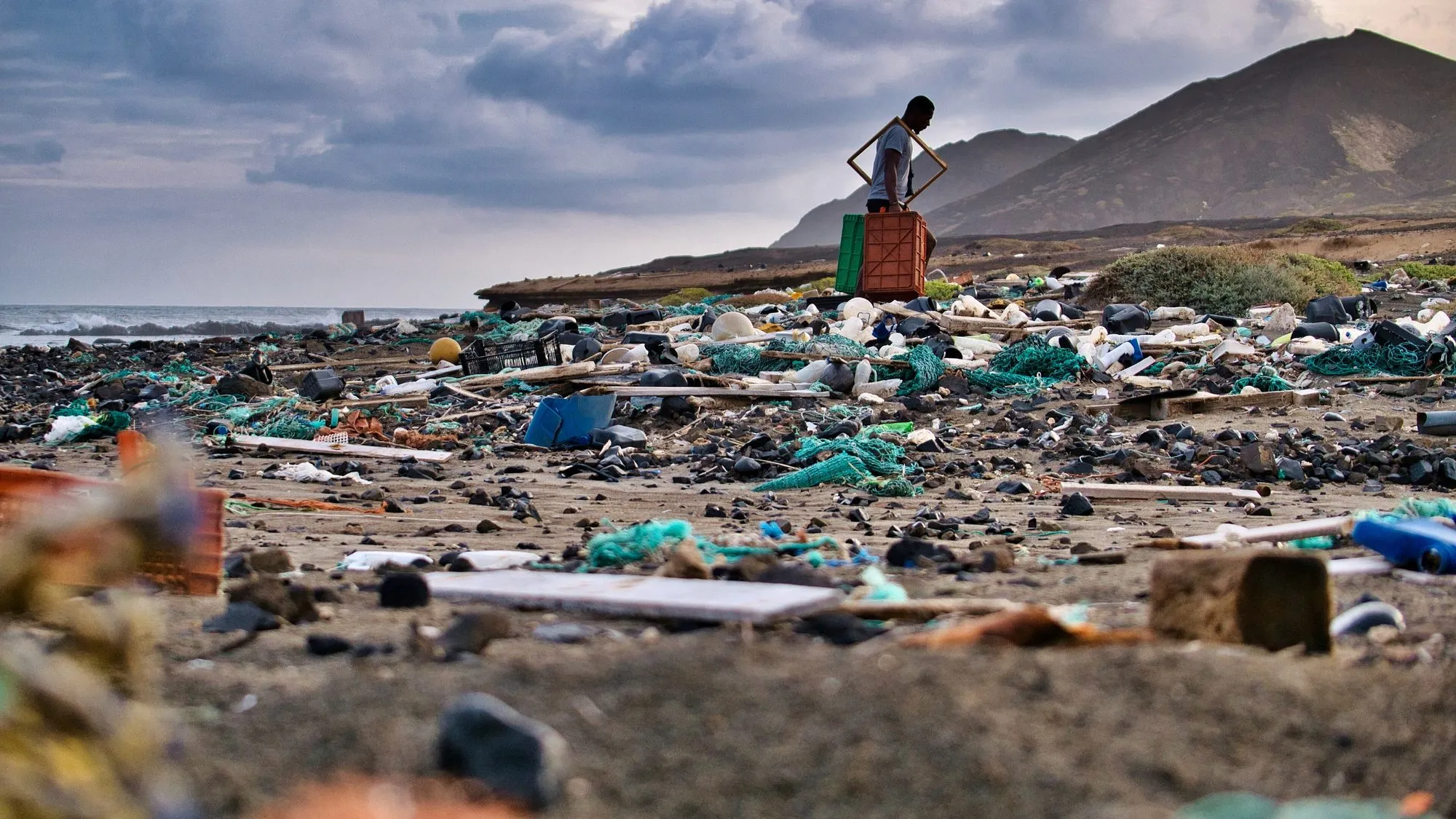 A beach strewn with plastic nets, buoys, bottles and boxes beneath a bare volcanic mountain, with a lone figure standing on the trash.