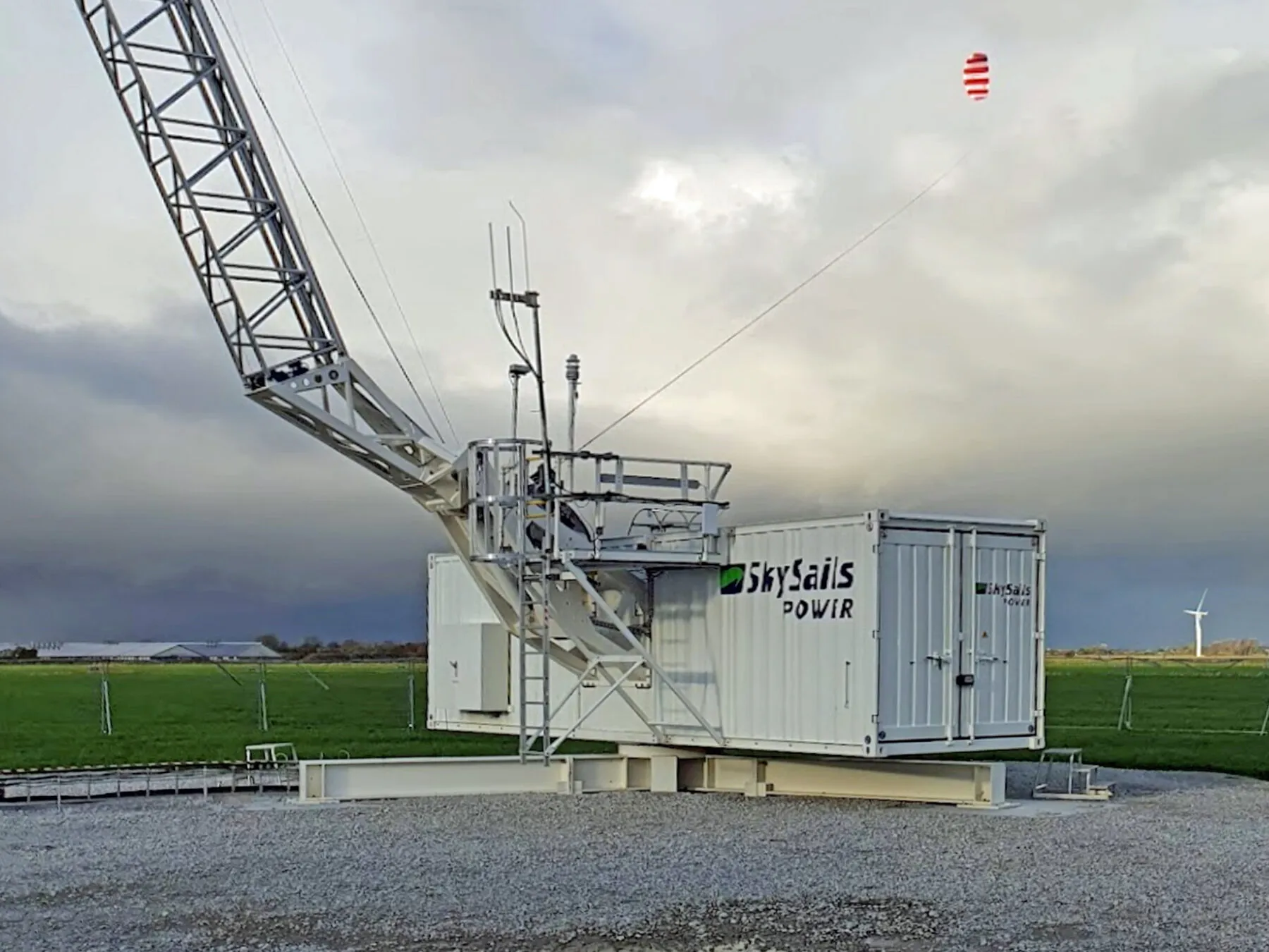 A SkySails Power ground station at Klixbüll: a white container with a tall lattice launch mast; far away on its tether a red and white kite flies in a grey sky above green fields.