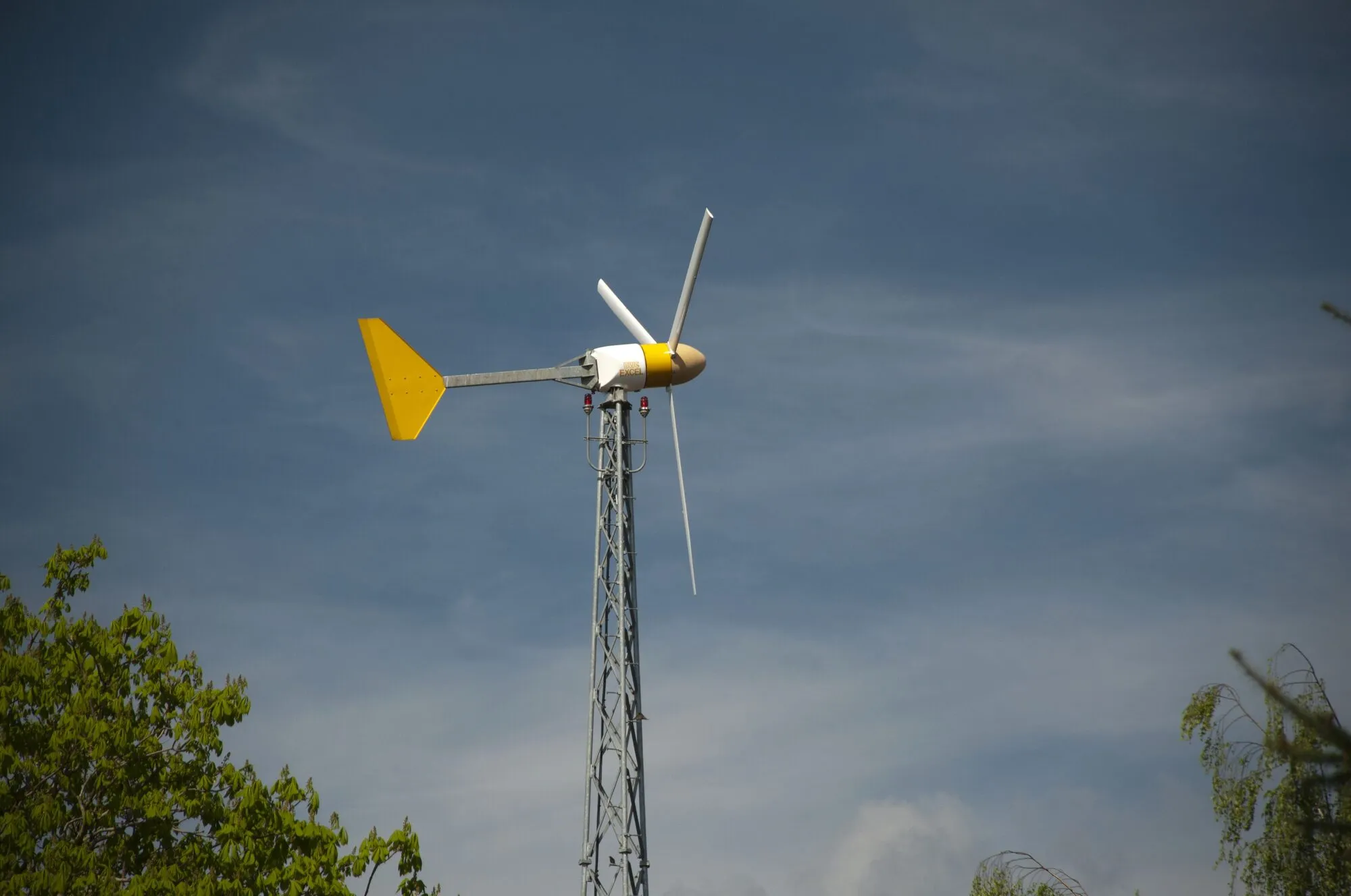 A small three-bladed wind turbine with a yellow tail vane on top of a tall lattice tower, against a blue sky above trees.