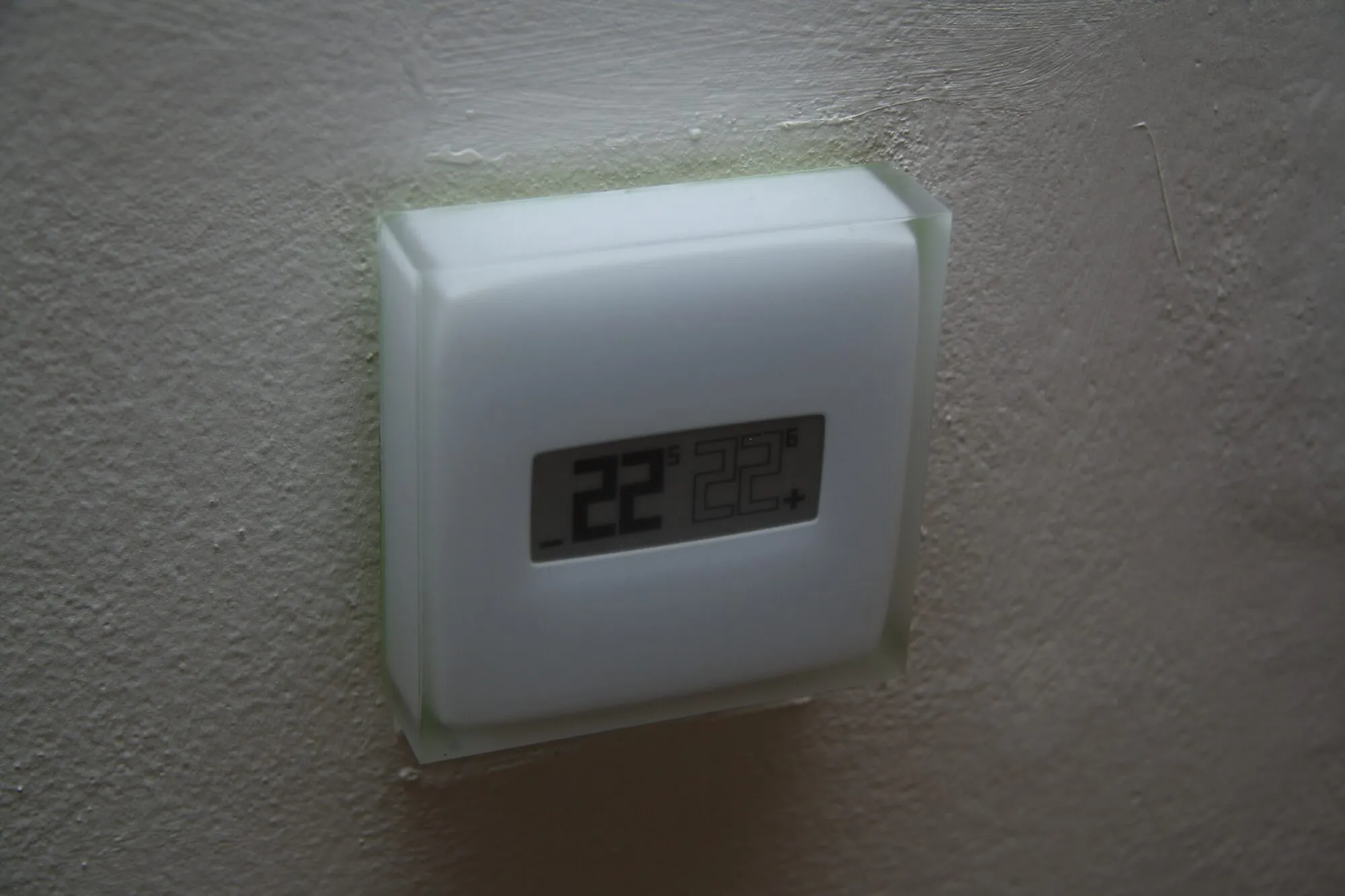 A small white smart thermostat with a digital display showing 22 degrees, on a plain wall.