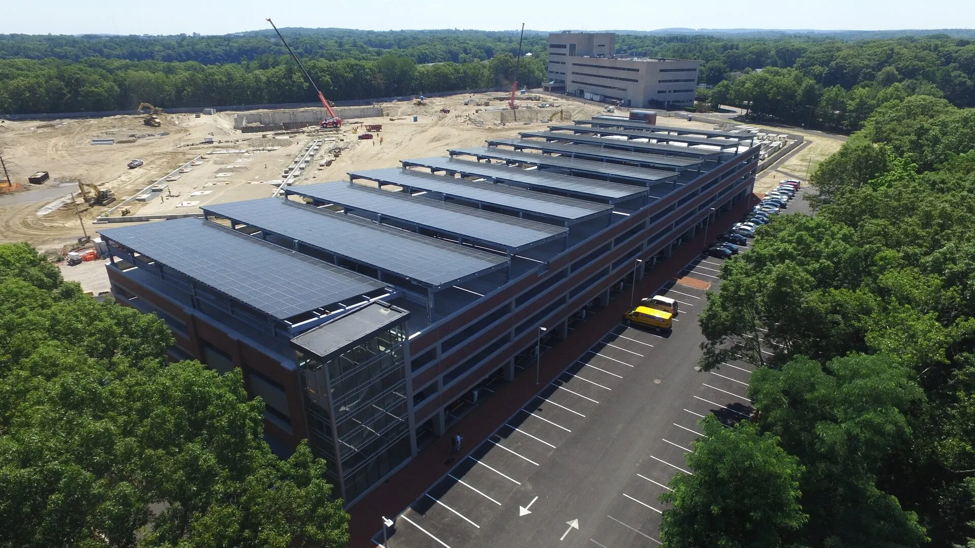 'n Luchtfoto van 'n parkeerterrein met verskeie verdiepings waarvan die boonste vloer bedek is met lang rye sonpanele op staal-balke, tussen bome.
