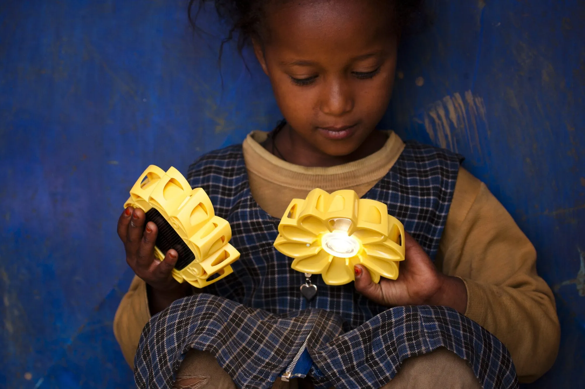 A young girl in a school pinafore looks down at a small yellow sunflower-shaped solar lamp burning in her hands.