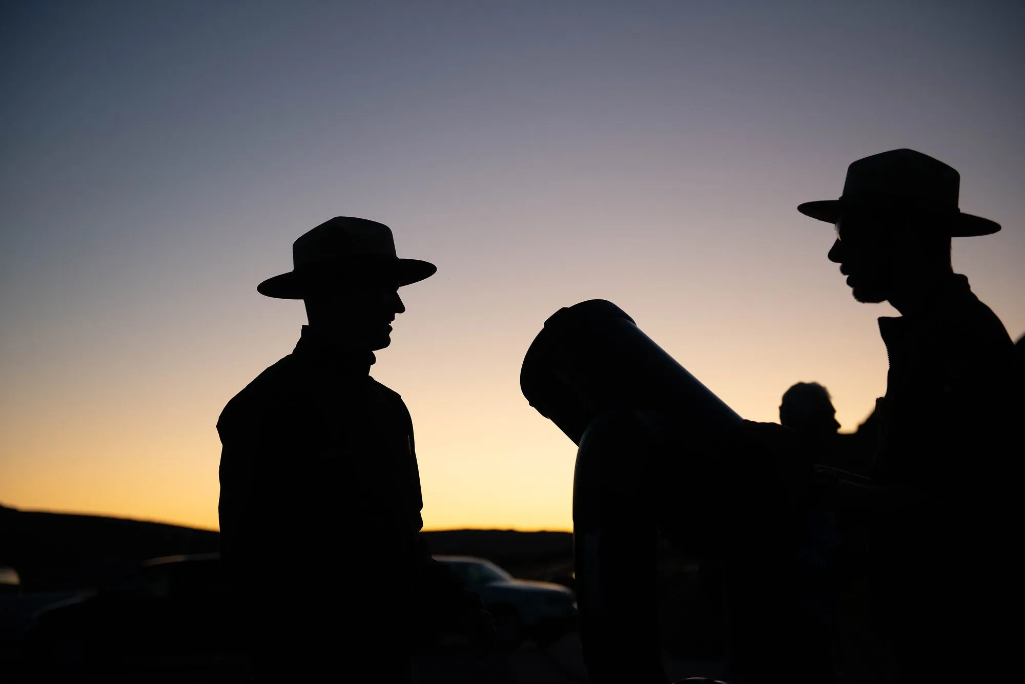 Twee park-rangers in silhouet naby 'n groot teleskoop teen 'n oranje middaghemel.