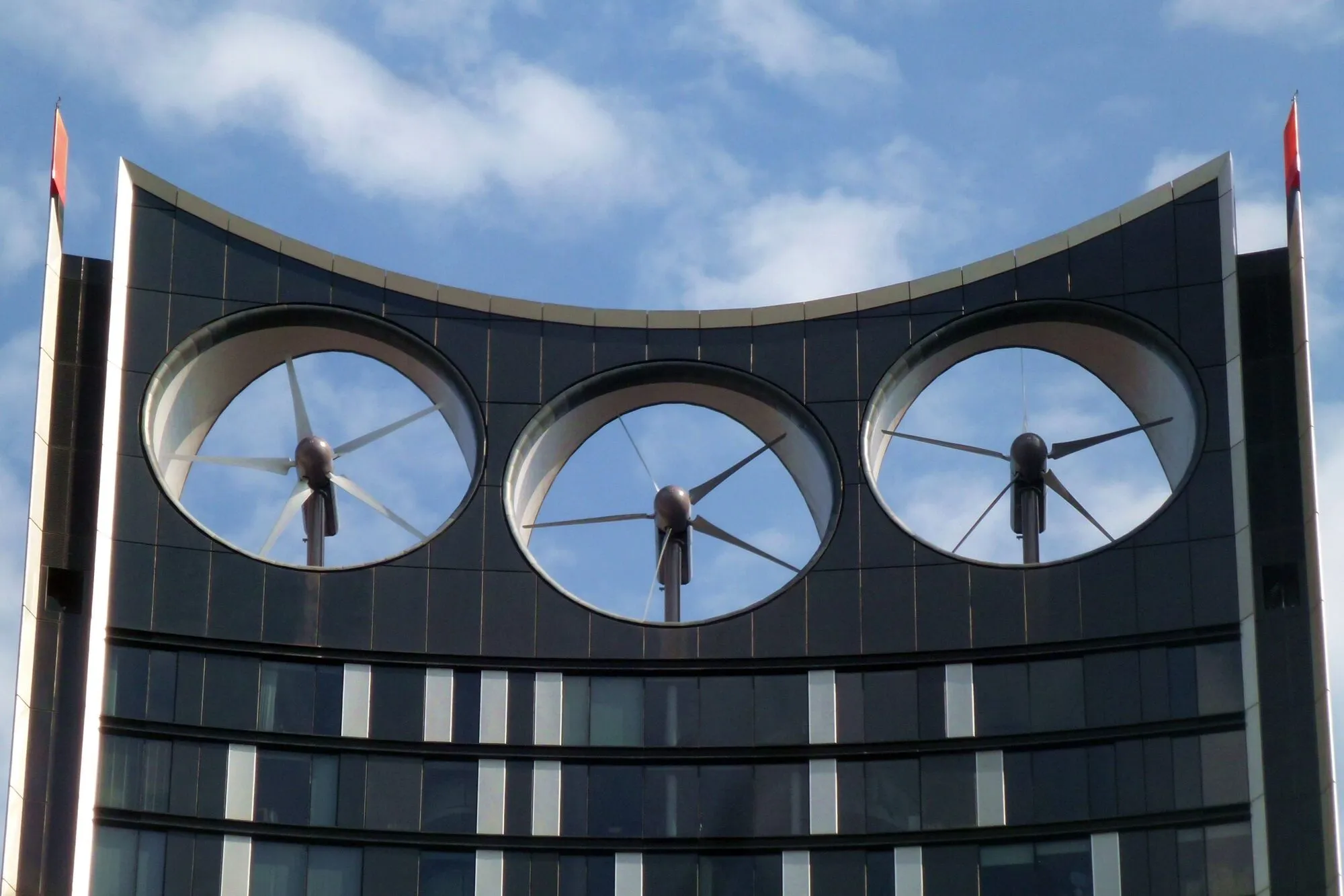 Three round openings in the curved top of a dark glass tower, each holding a wind turbine rotor, against a blue sky.