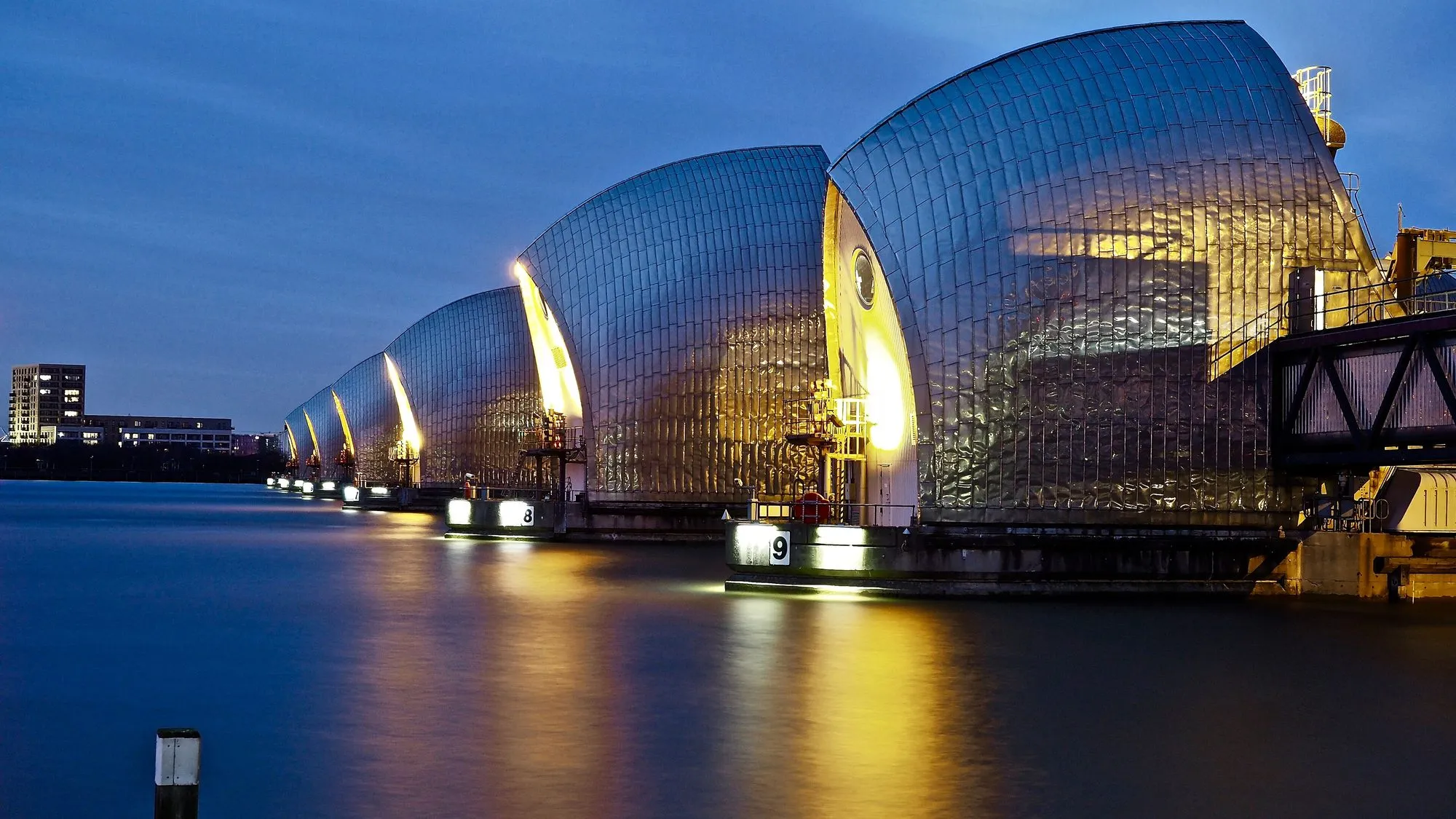 Die glansende staalkappe van die Thames Barrier-piere verlig in die skemer, weerspieël in die kalme rivier.