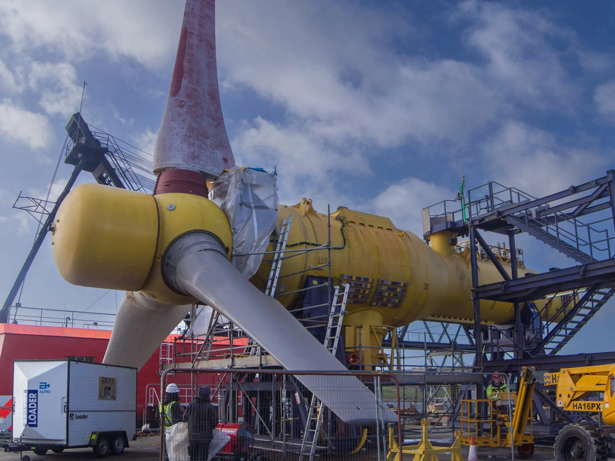 A large yellow tidal turbine with three pale blades on a quayside, surrounded by scaffolding, a mobile platform and workers.