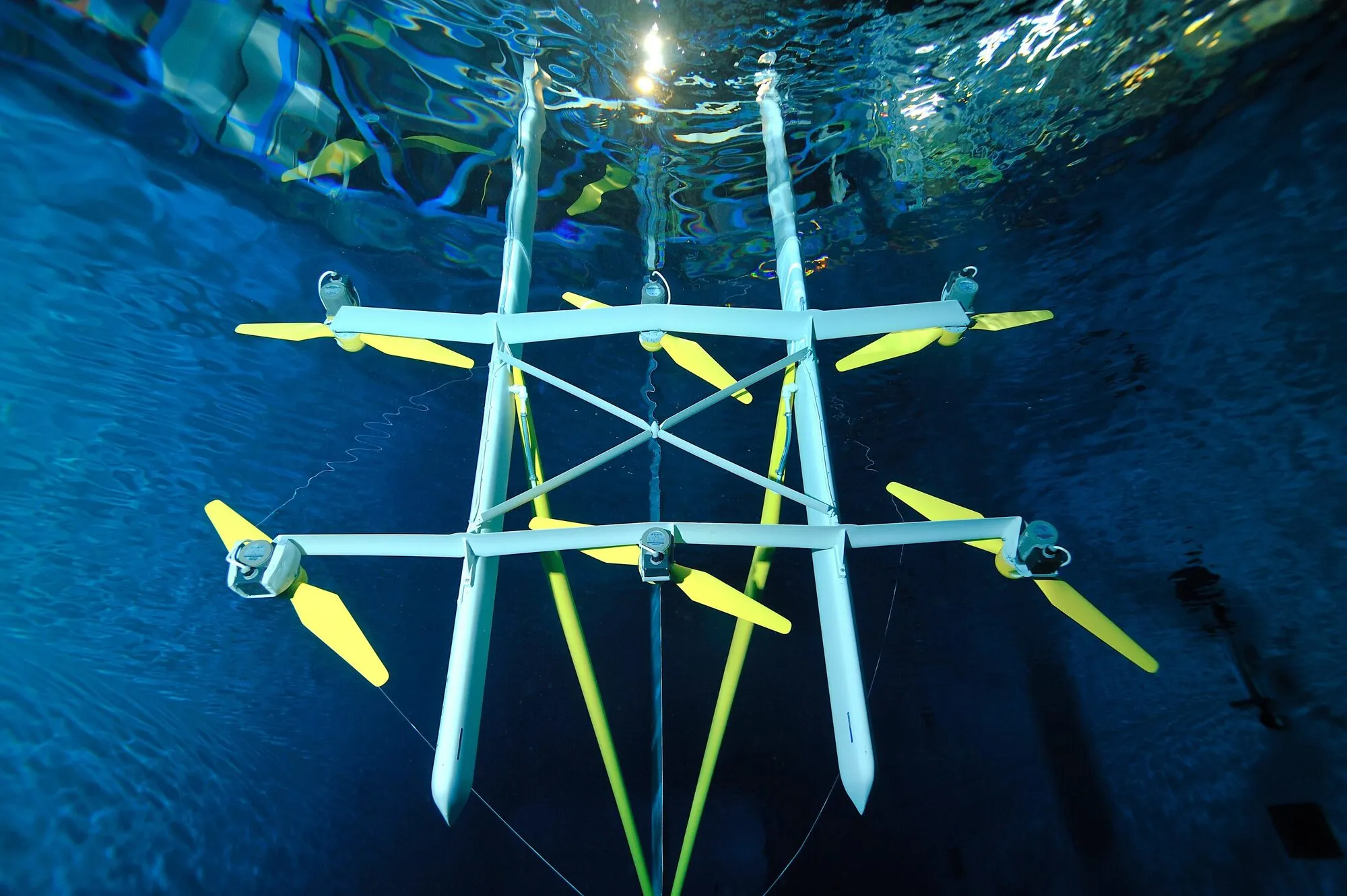 Underwater view of a model tidal turbine with several yellow two-bladed rotors on a light blue frame in a test basin.