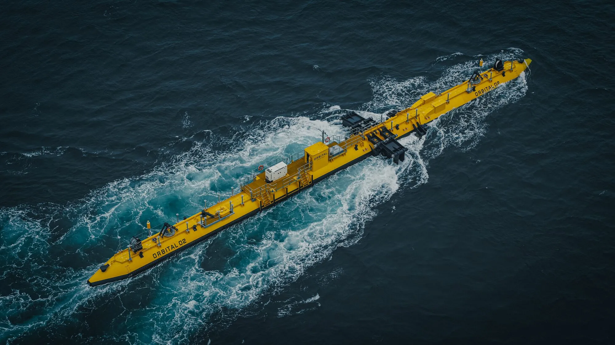 Aerial view of a long yellow floating tidal turbine in a fast current, with white water streaming past its hull.