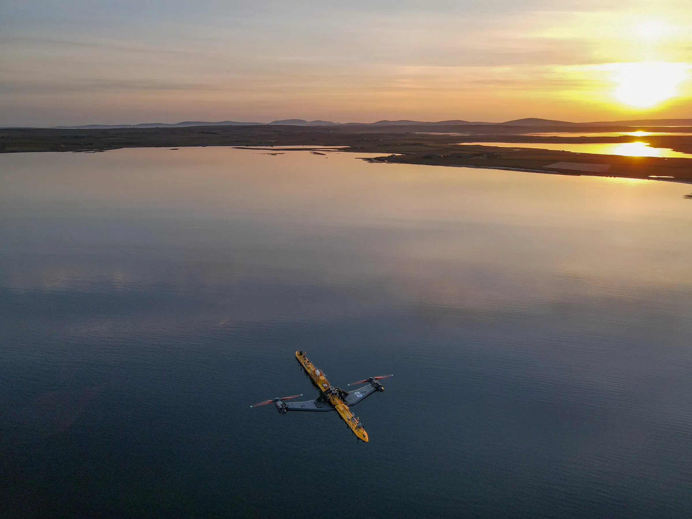 A long yellow floating tidal turbine moored on a calm sea at sunset, with low islands on the horizon.