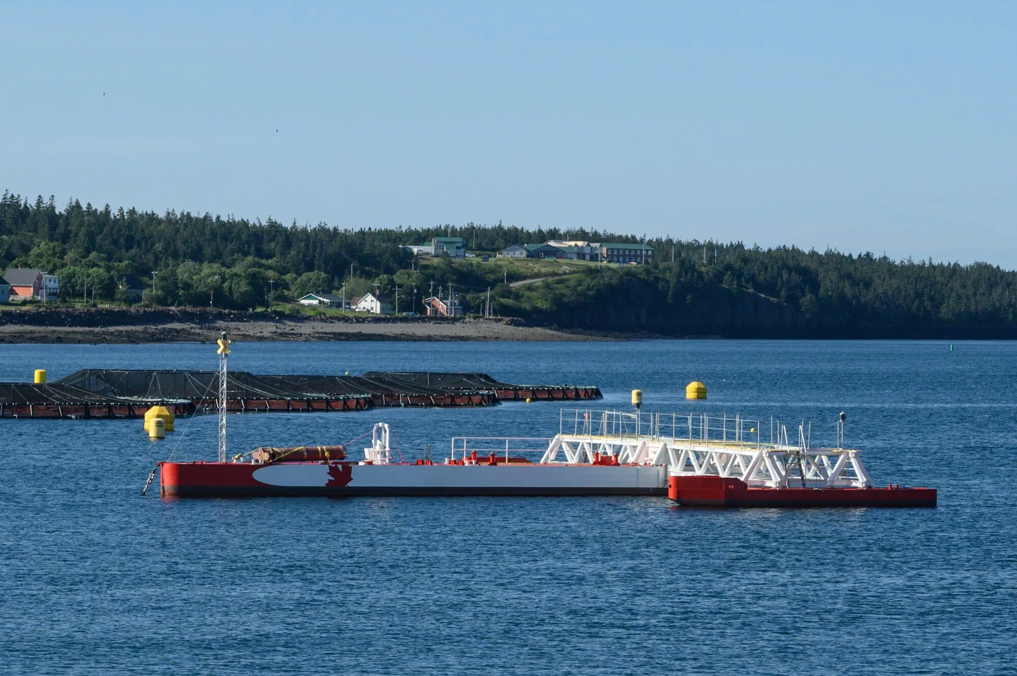 A red and white floating platform with tidal turbines moored in a calm harbour channel, with a wooded shore behind.