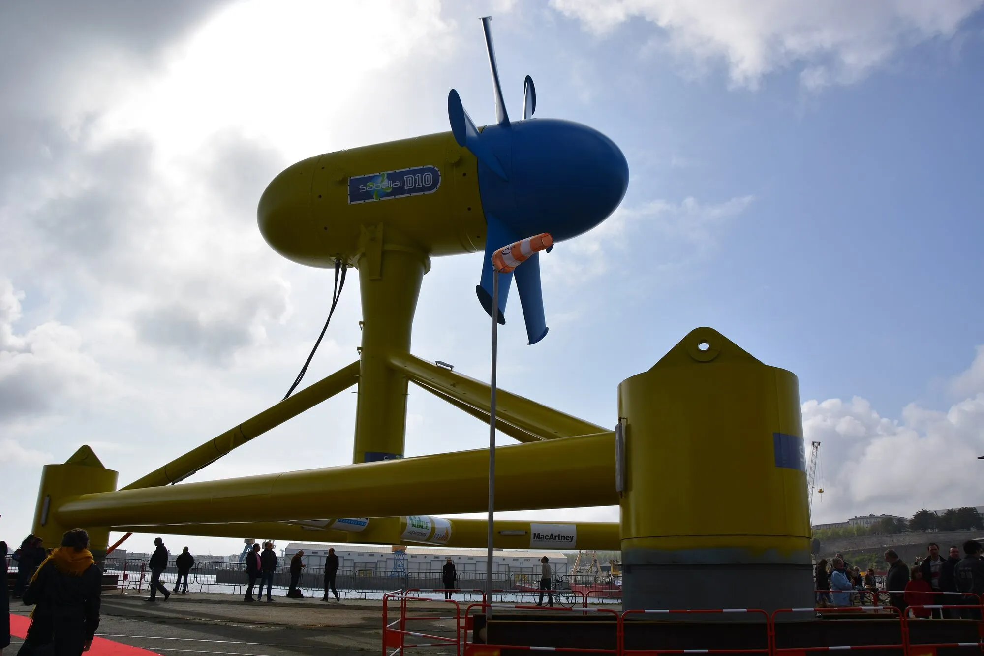 A large yellow tidal turbine with a blue nacelle and rotor, standing on its tripod base on a quayside with visitors around it.
