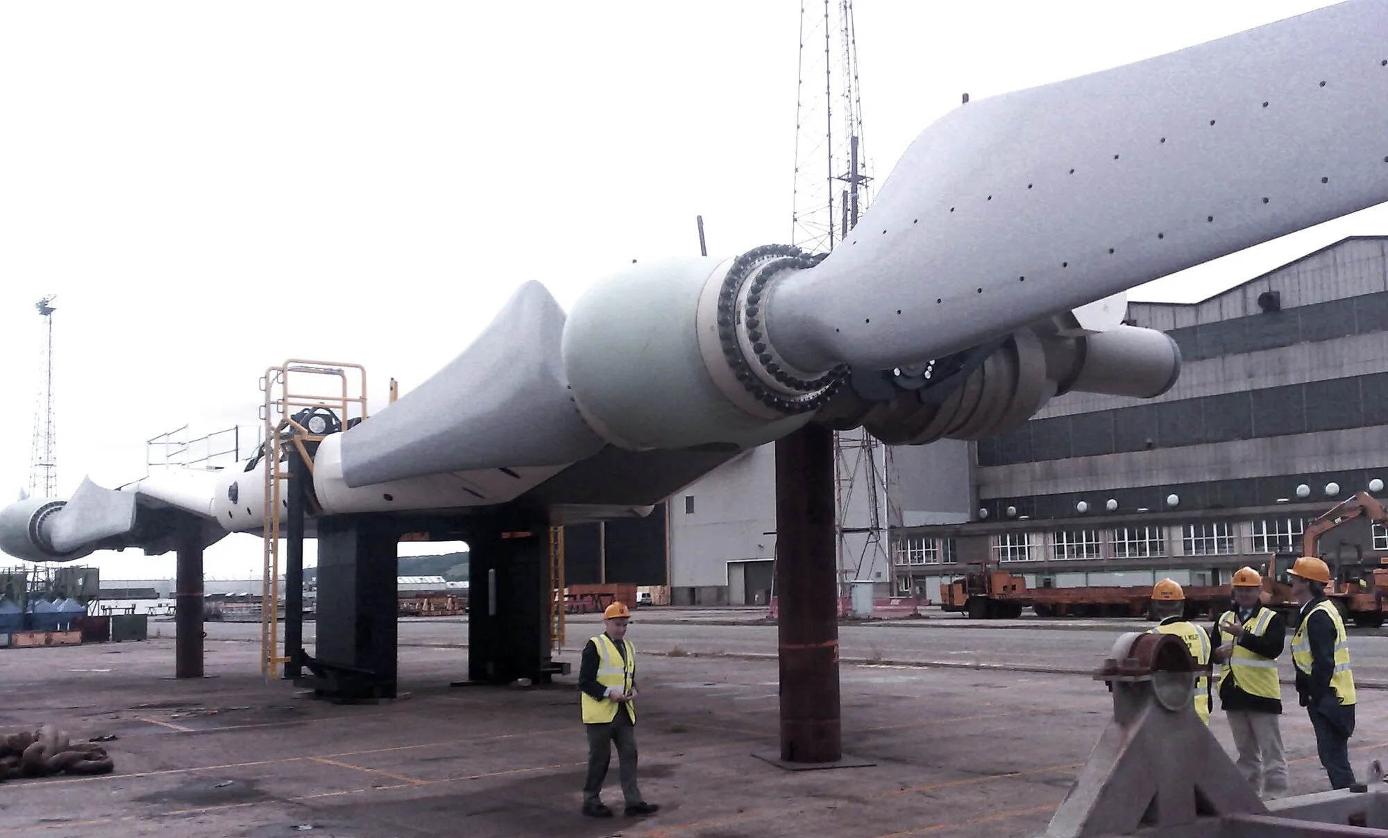 Two long grey turbine rotors on a steel frame on a shipyard quay, with workers in high-visibility jackets beside them for scale.
