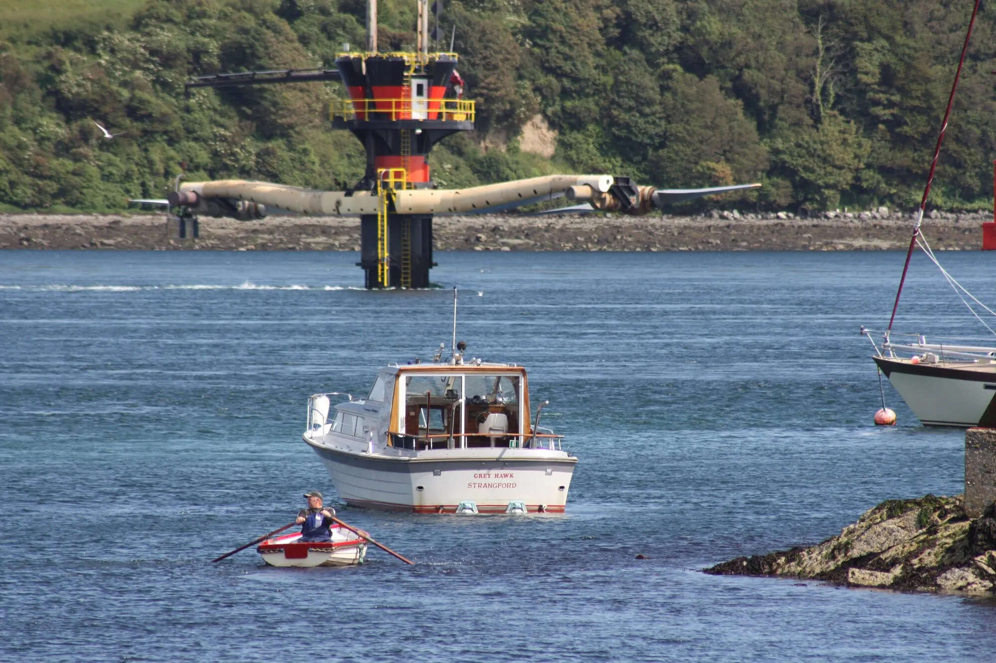 A tidal turbine on a pile in a narrow sea channel, its two rotor arms raised clear of the water, with a small boat passing in front.