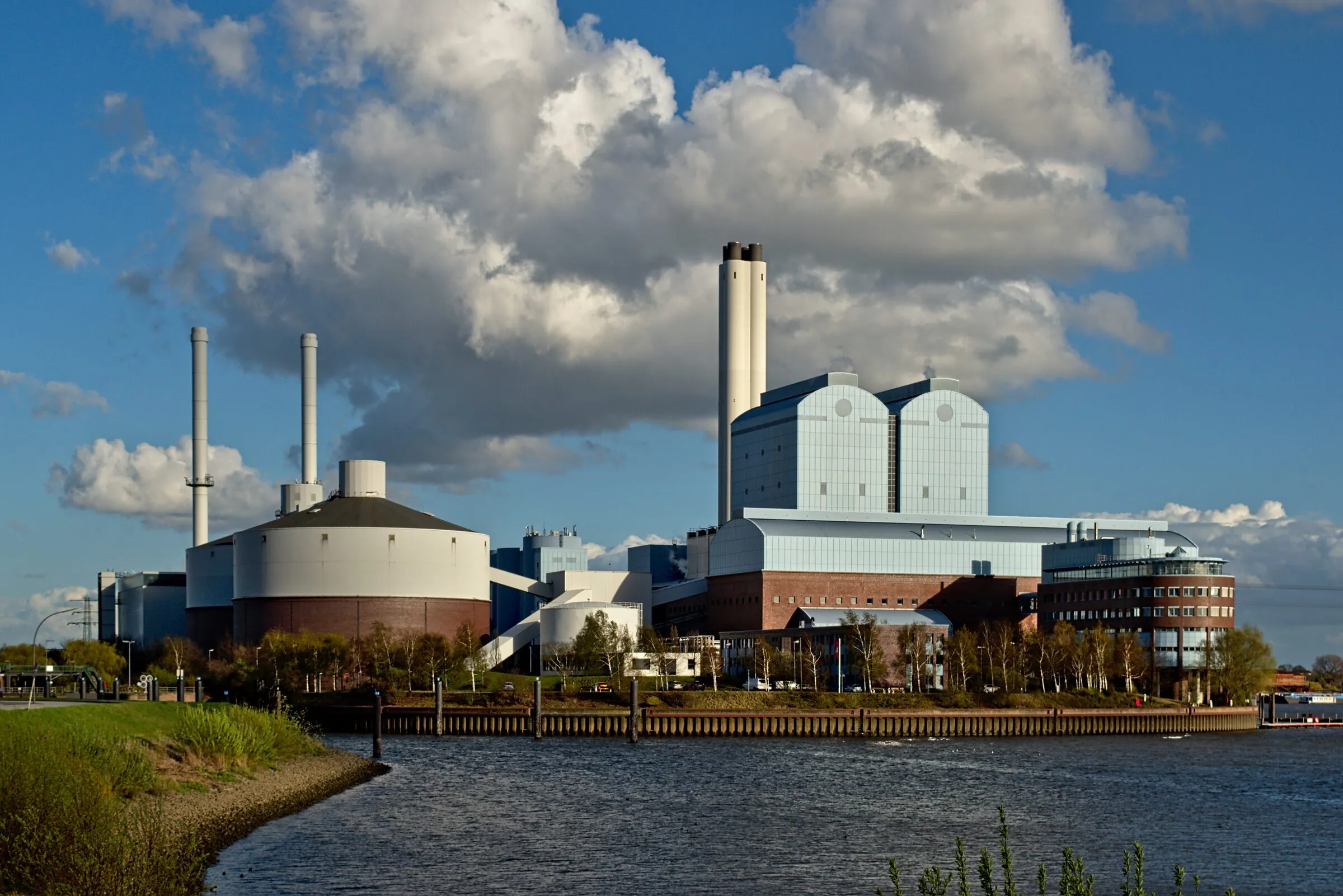 A district heating station with tall chimneys beside a river in Hamburg, the kind of network a heat battery feeds.