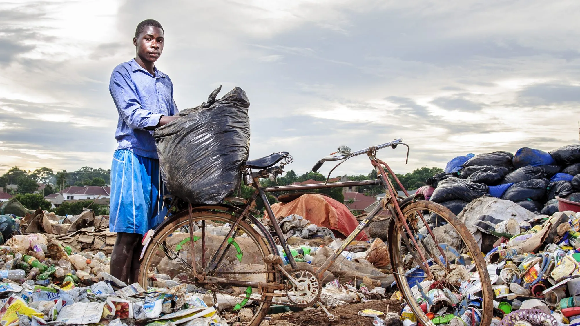 A young man with a bicycle loaded with a large black bag of collected plastic stands on a trash dump under a cloudy sky.