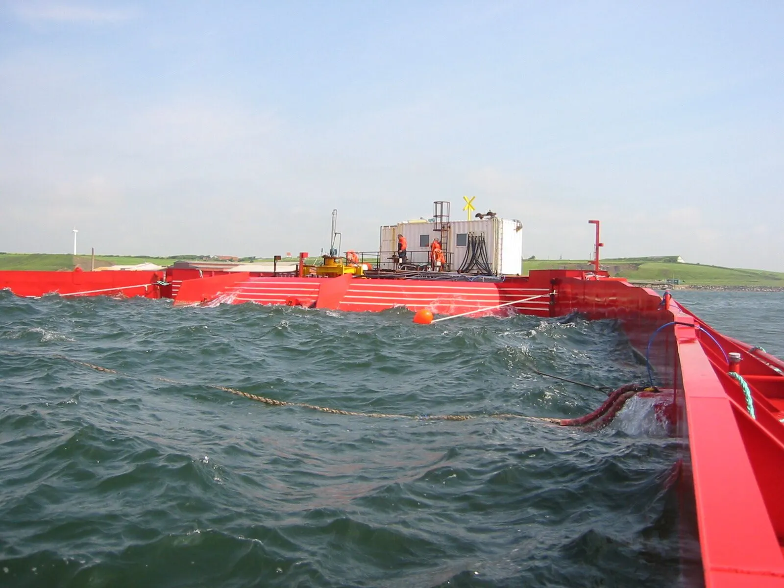 A large red floating structure with two long arms and a ramp, moored in a grey sea near a green shore.