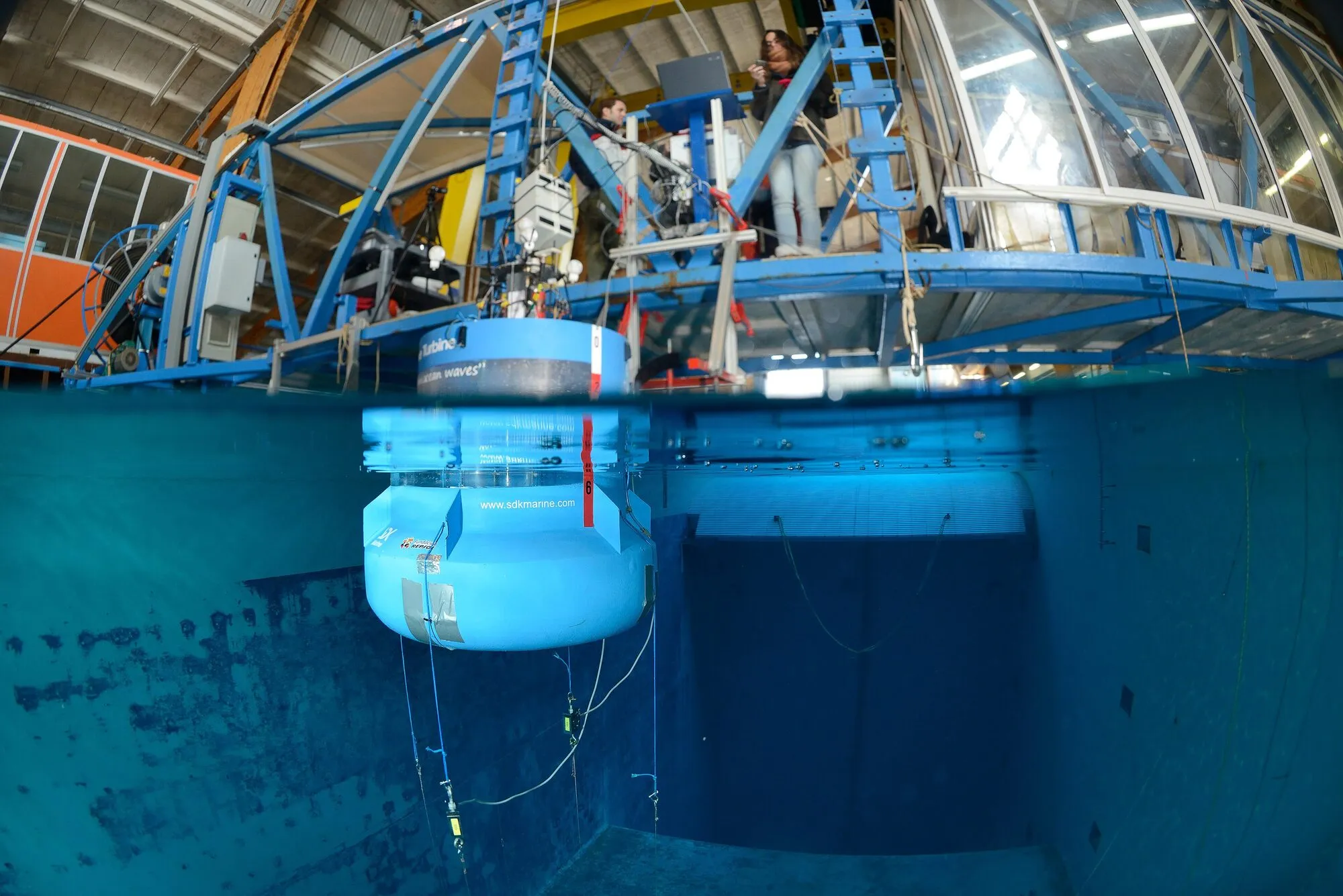 Half above and half below the water line of a test basin: a model wave energy buoy hangs from a blue steel gantry with instruments.