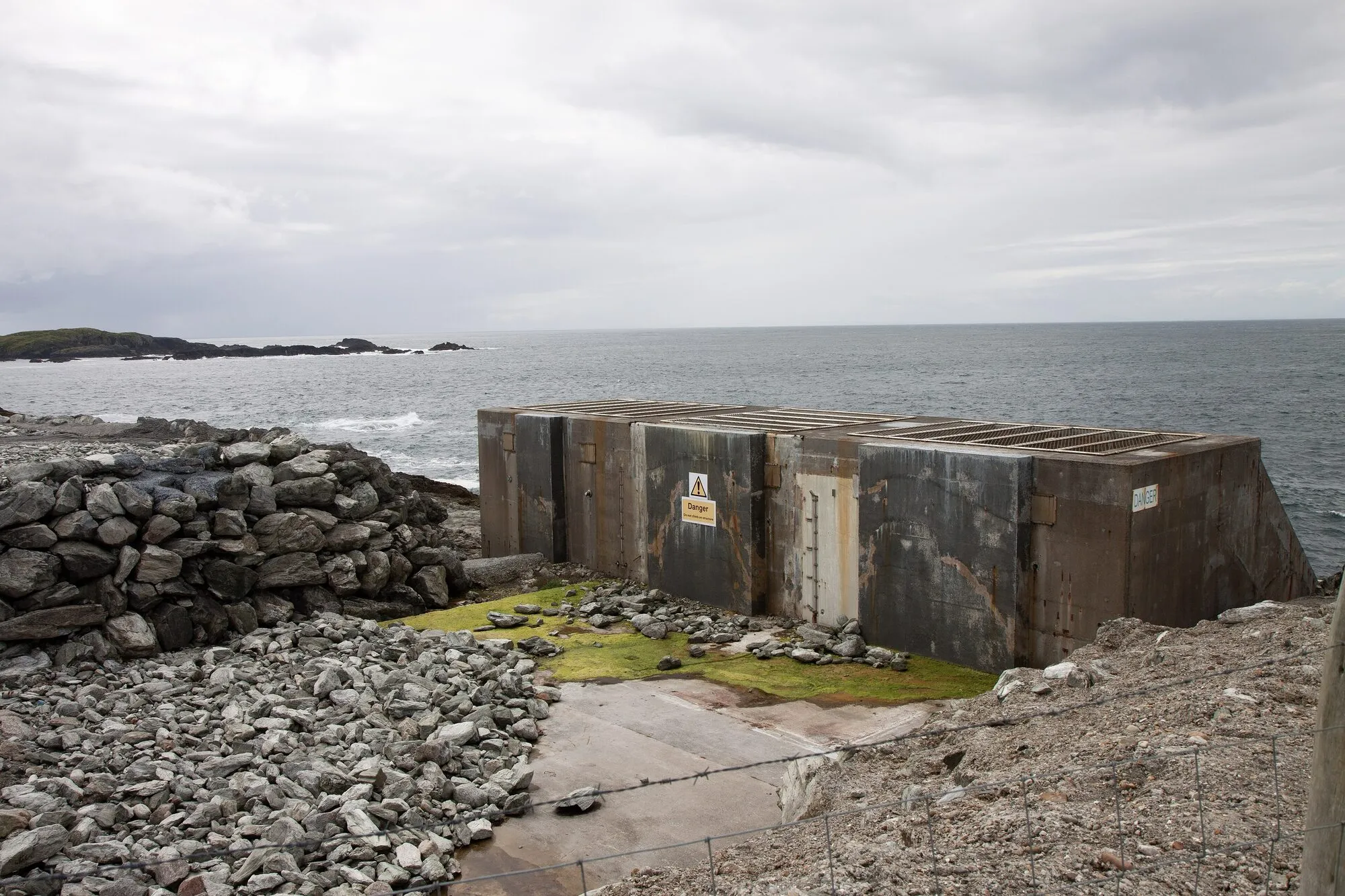 A roofless concrete chamber on a rocky shoreline beside the sea, the remains of a wave power station.