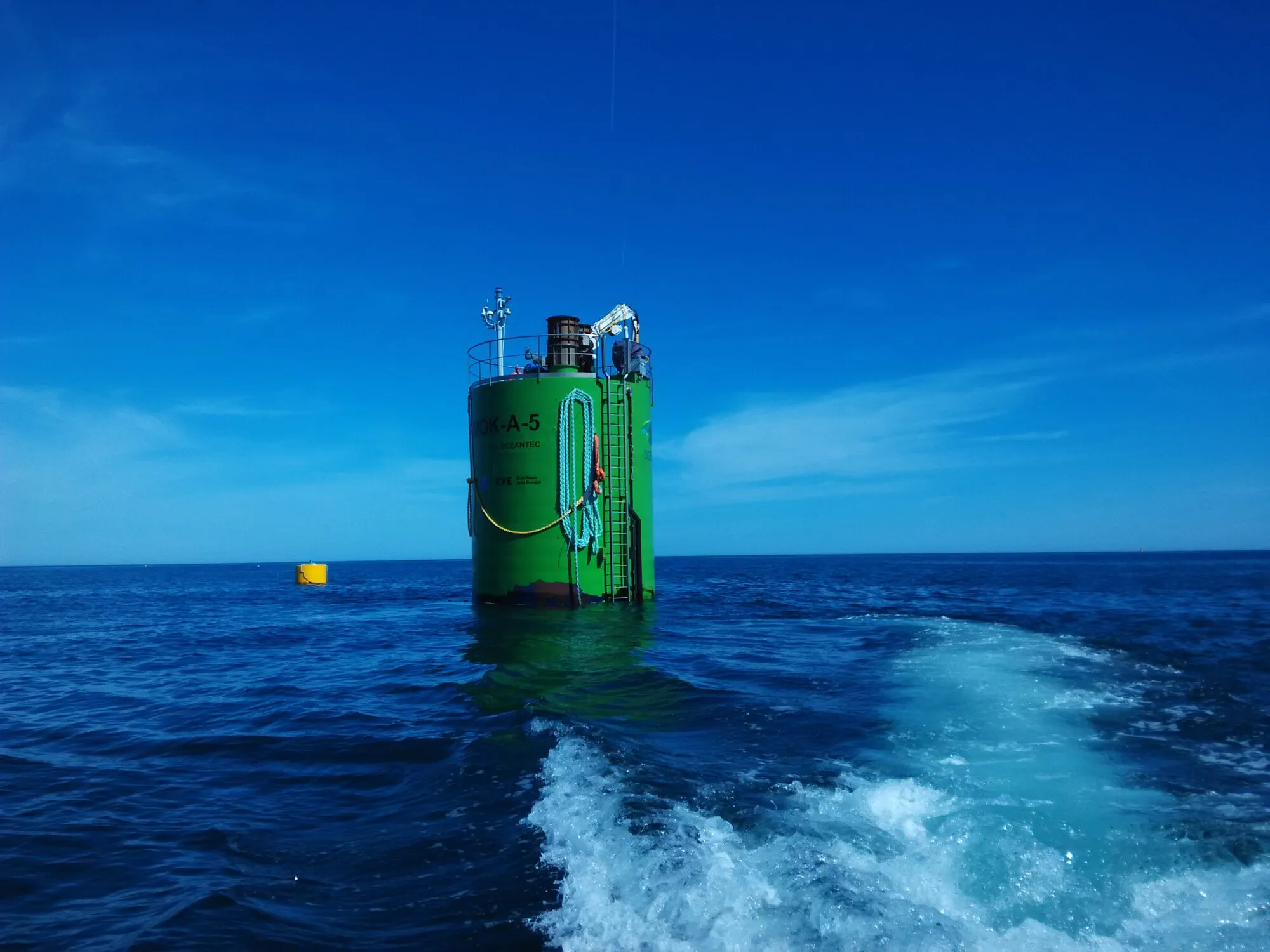 A green cylindrical buoy floating upright in a deep blue sea, seen from the wake of a boat.