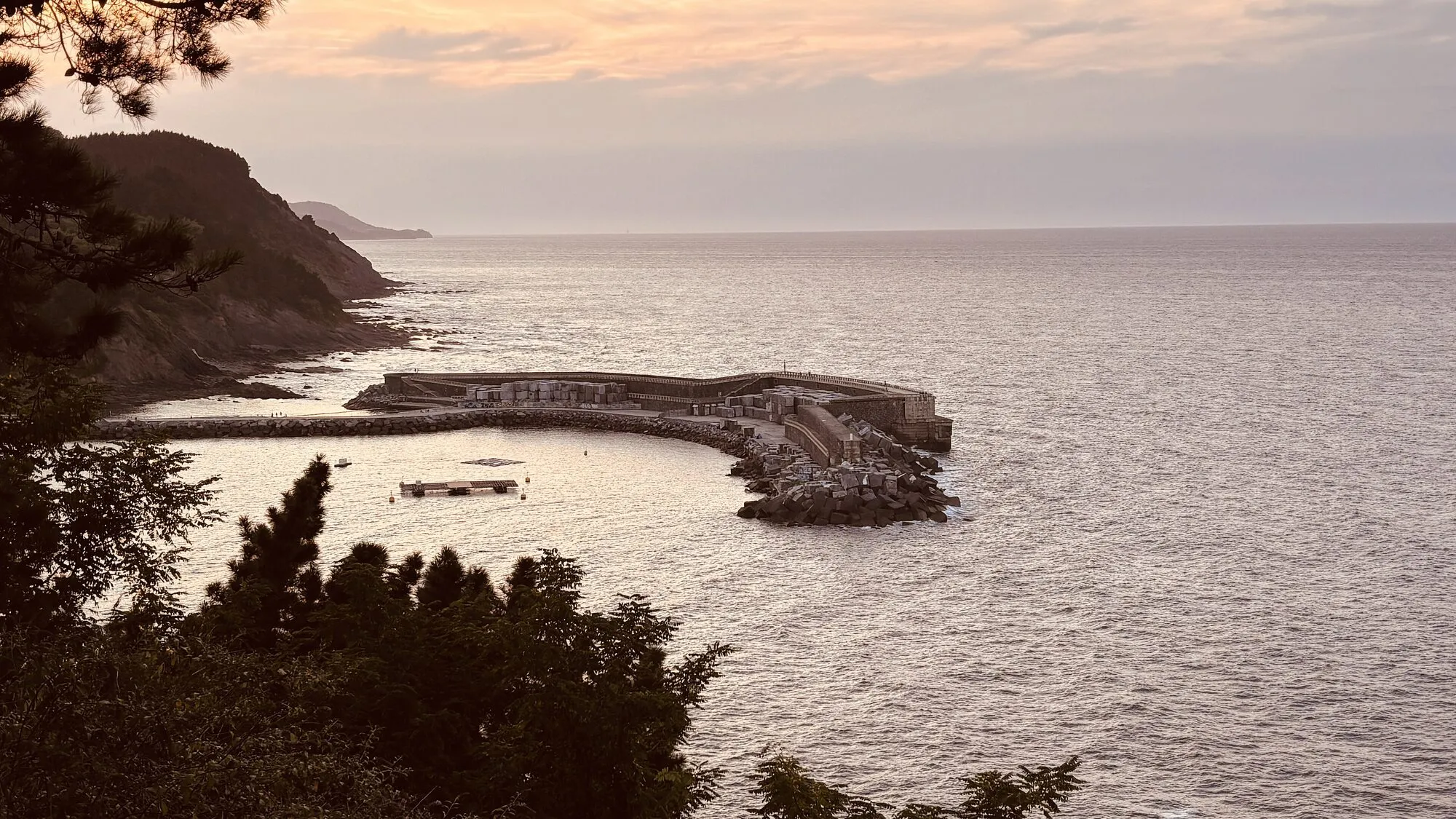 A curved harbour breakwater running out into a calm sea at dusk, below a wooded headland.