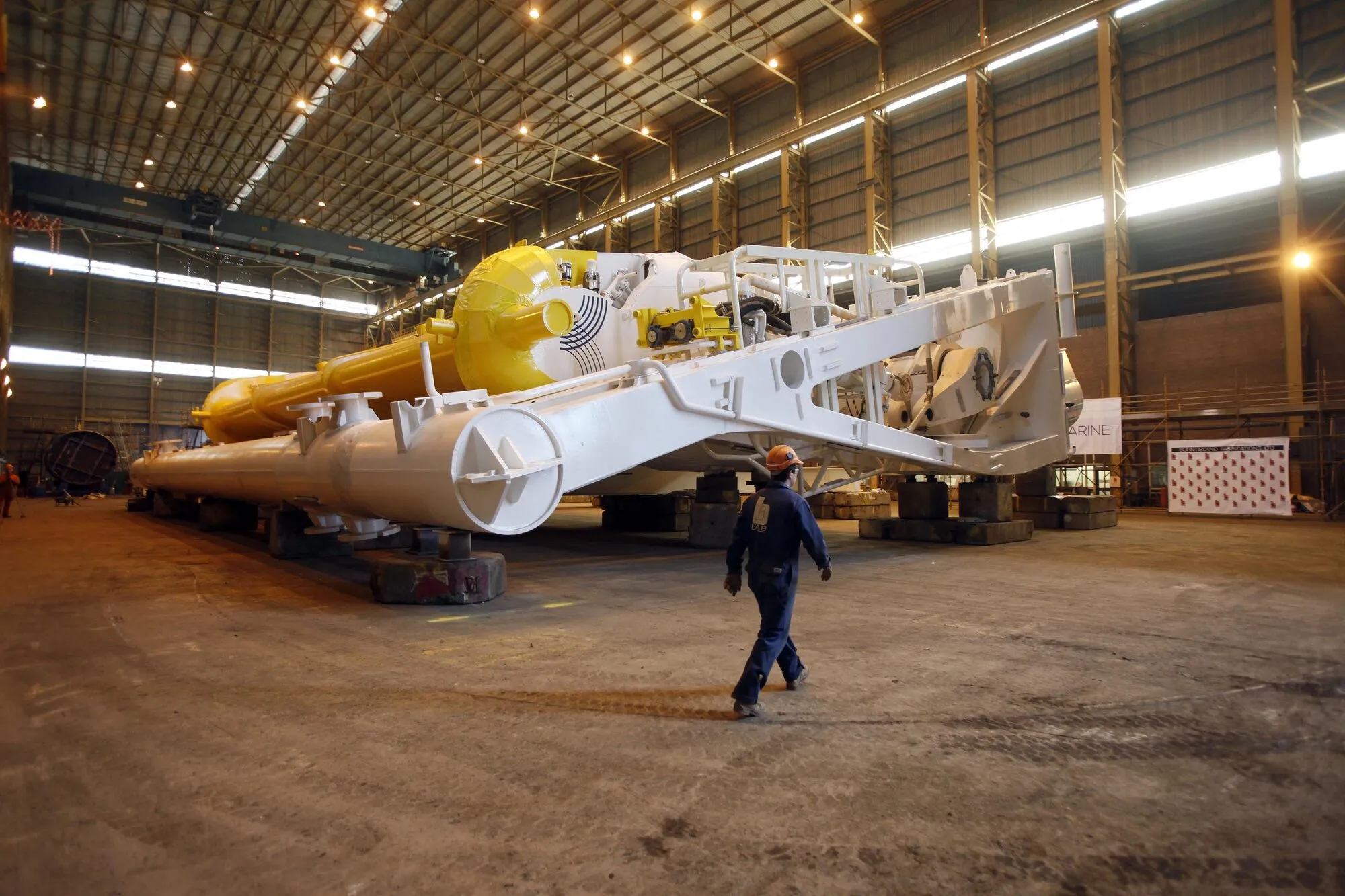 A large yellow and white hinged steel flap lying in a fabrication hall, with a worker walking past it for scale.
