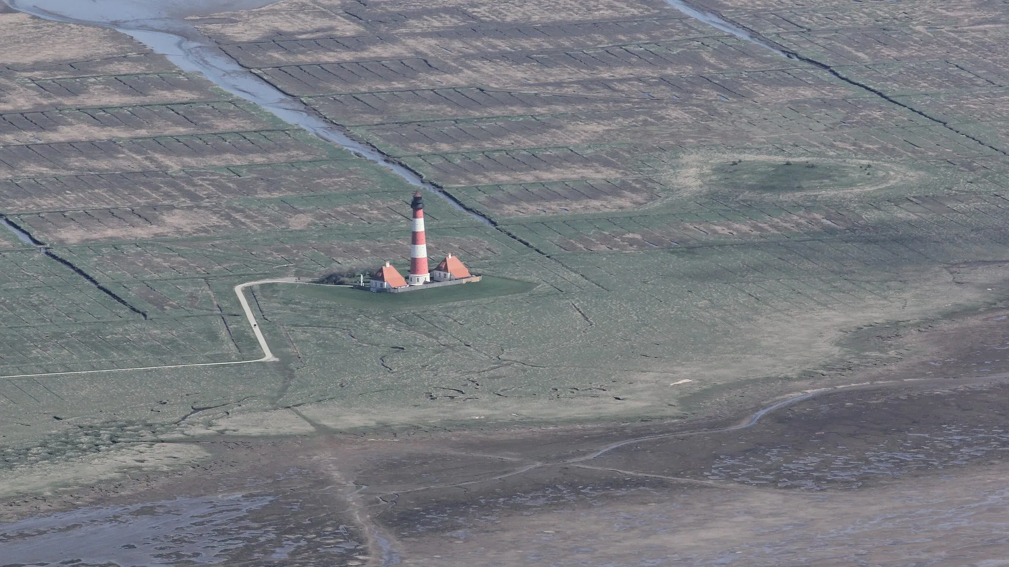 Lugfoto van 'n rooi en wit vuurtoring wat in 'n wye groen zoutmoerasse staan, gesny deur krewe, aan die rand van gety-moddervlaktes.