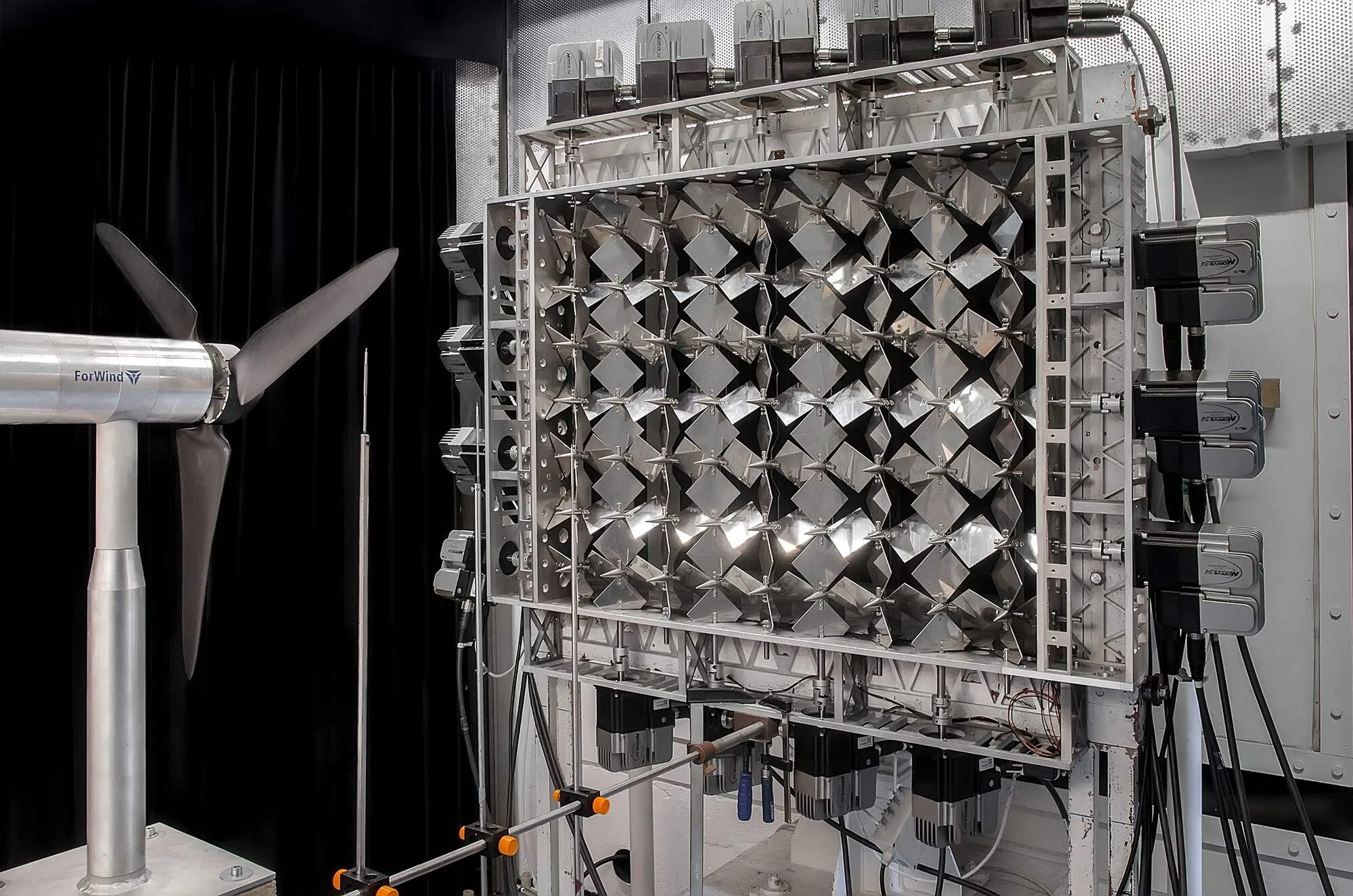 A small three-bladed model wind turbine on a slim tower in a wind tunnel, in front of a large grid of metal flaps driven by motors.